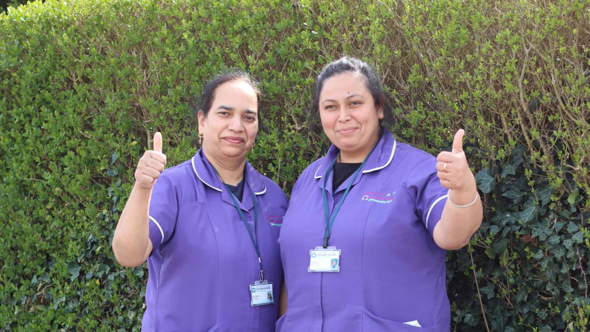Two smiling female healthcare workers in purple uniforms stand in front of a hedge, giving a thumbs up to the camera. Both are wearing ID badges and provide home care services in Slough.
