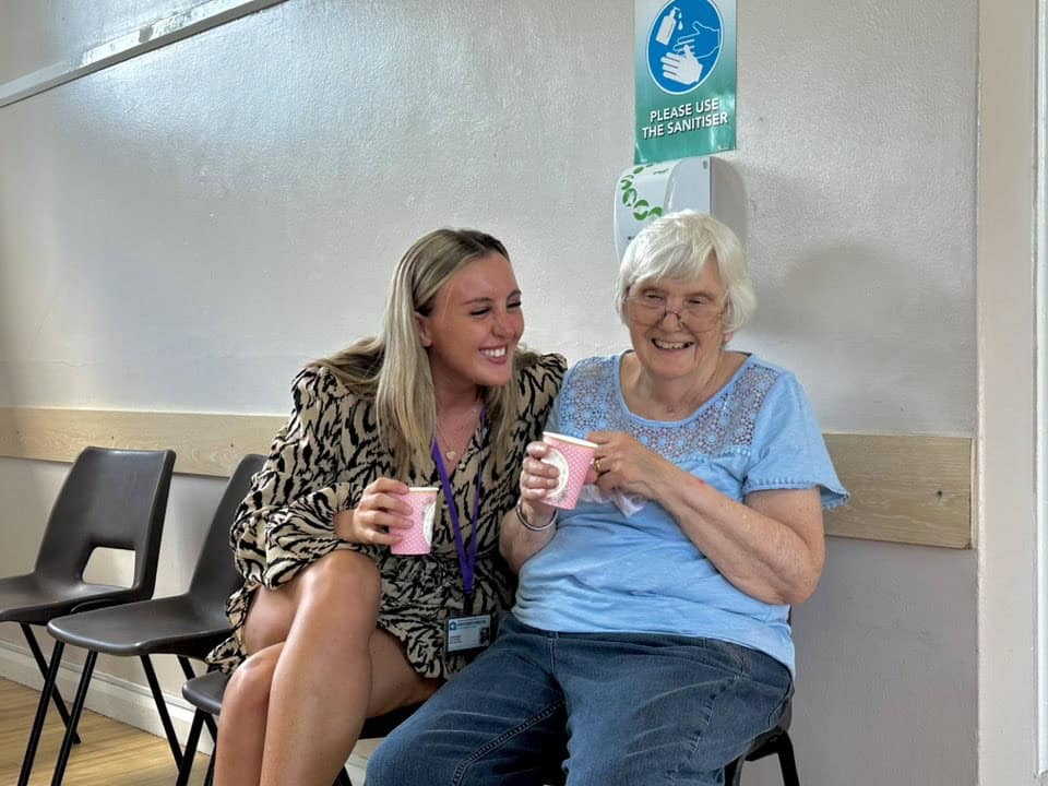 A young woman and an older woman sit closely together on chairs, smiling and holding pink disposable cups. A hand sanitizer sign is visible, highlighting the caring atmosphere often found in live-in care as they enjoy a cheerful moment together.