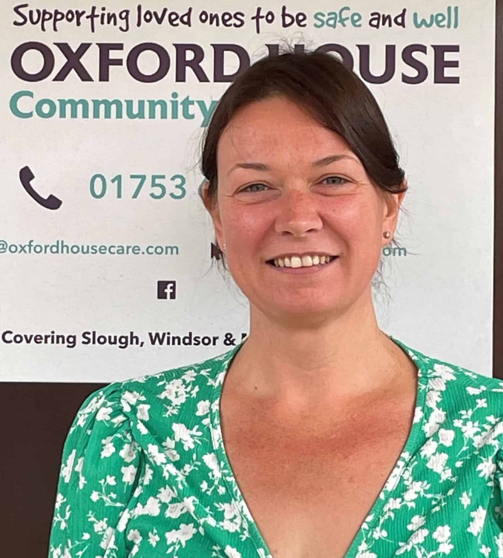 A smiling woman in a green floral top standing in front of a sign that reads "supporting oxford house community" with contact information and areas covered, including slough and windsor, indicated below.