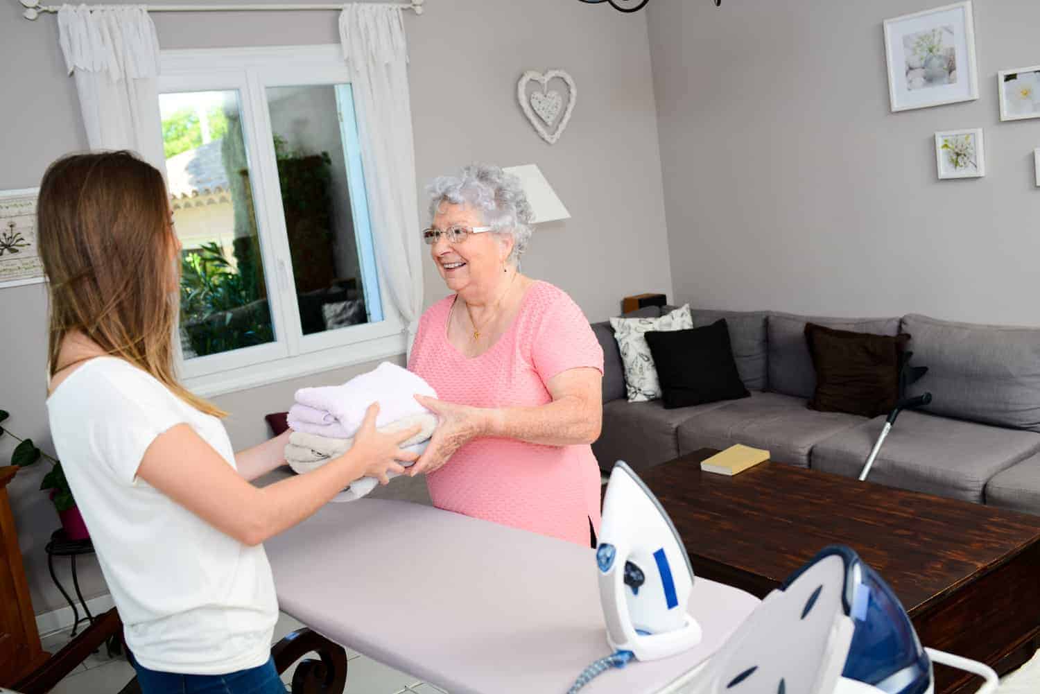 A smiling elderly woman receives a stack of freshly folded towels from a carer in a homey living room, with an ironing board and iron in the foreground
