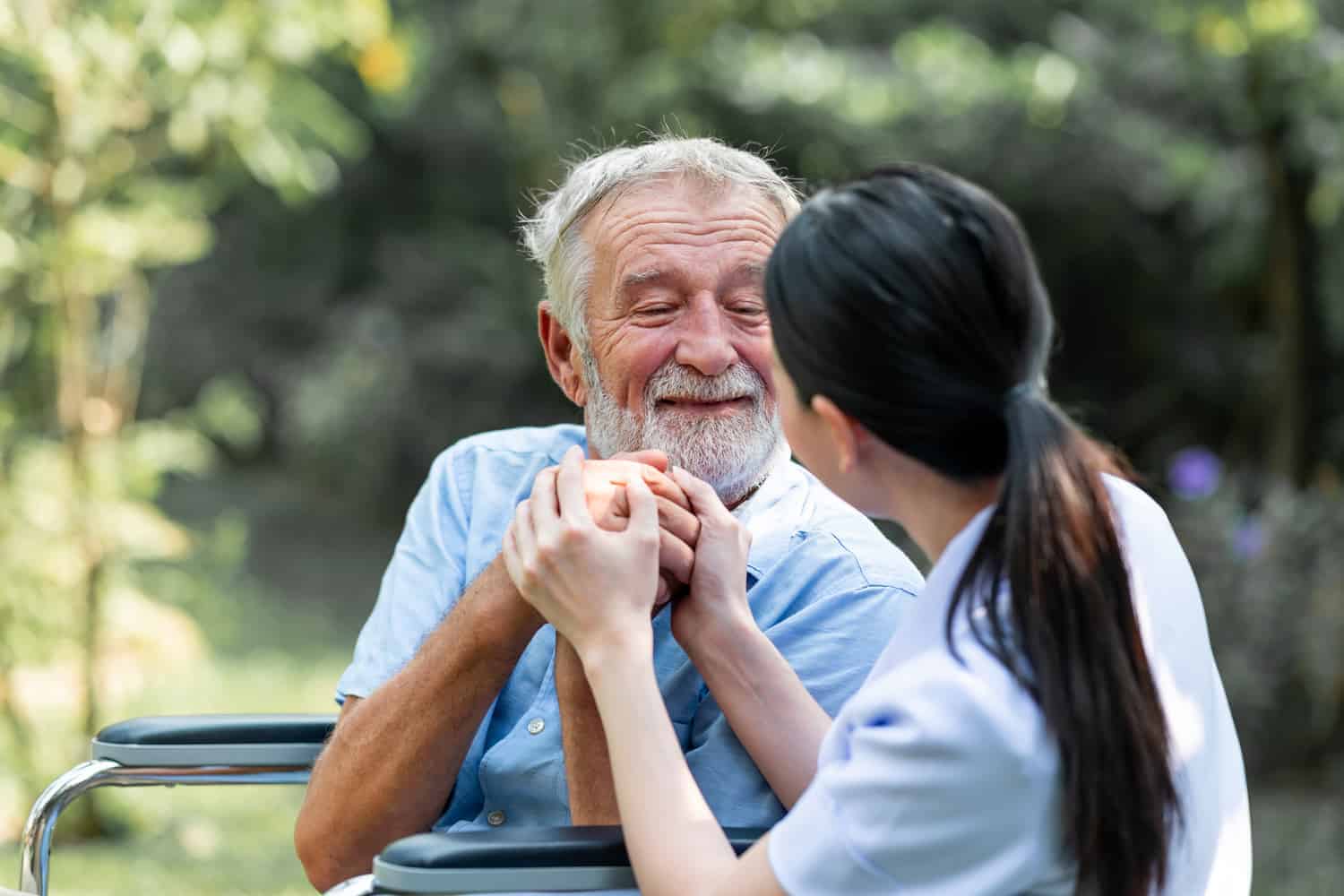 A tender moment in daily life help: a caregiver and an elderly man in a wheelchair, sharing a warm and comforting hand-hold in a lush park setting.