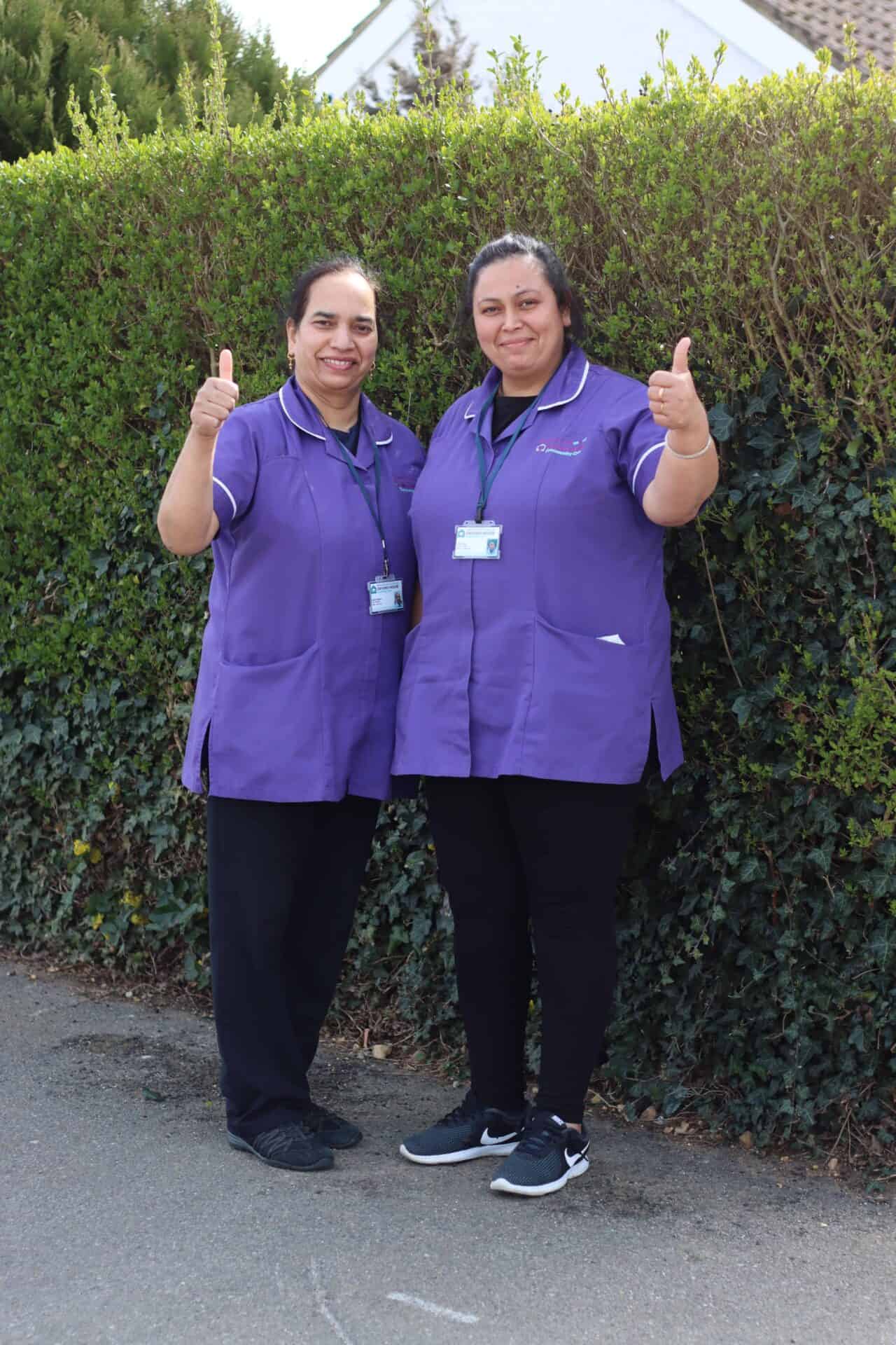 Two smiling healthcare professionals in purple scrubs giving a thumbs-up outside.