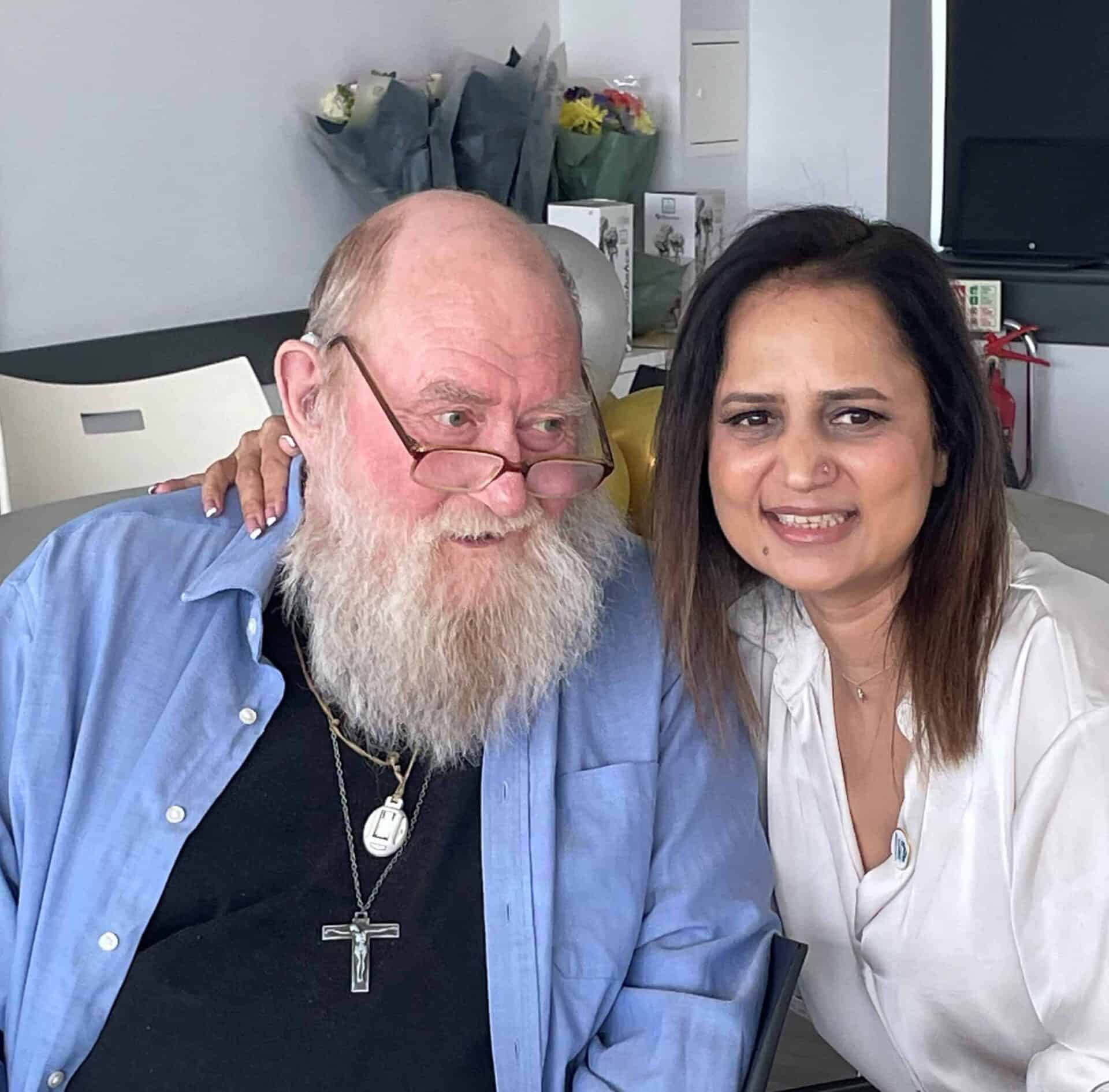 Two smiling individuals posing together for a photo, with one person sporting a long white beard and glasses, and the other with shoulder-length hair. The caregiver and their companion stand in front of a homely