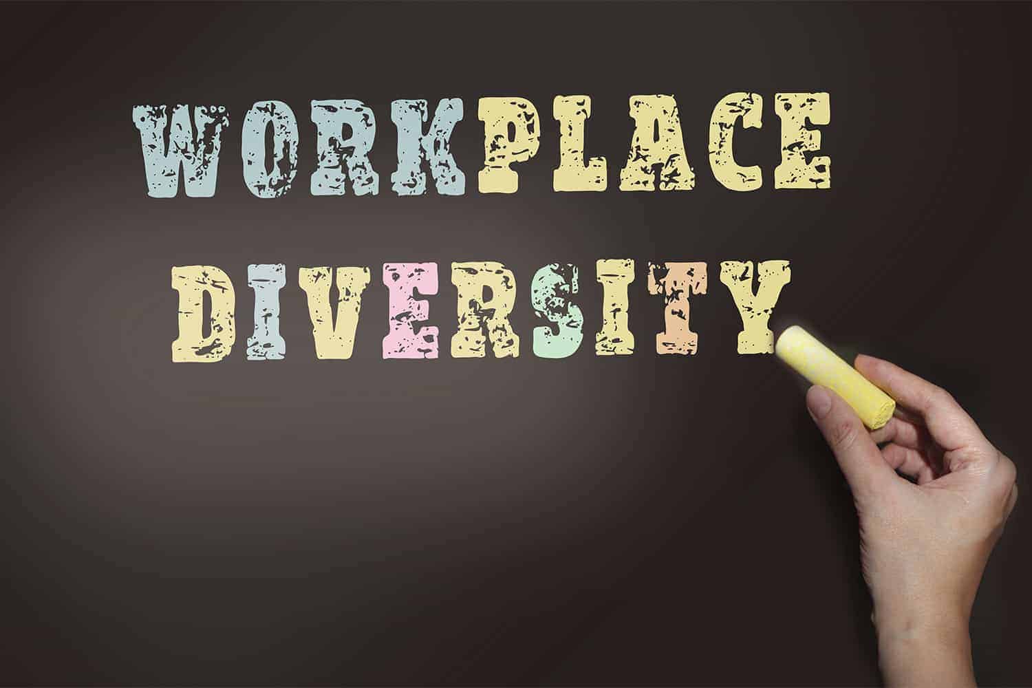 A hand holding a piece of chalk against a chalkboard, with the words "workplace diversity" written in colorful, distressed font style.