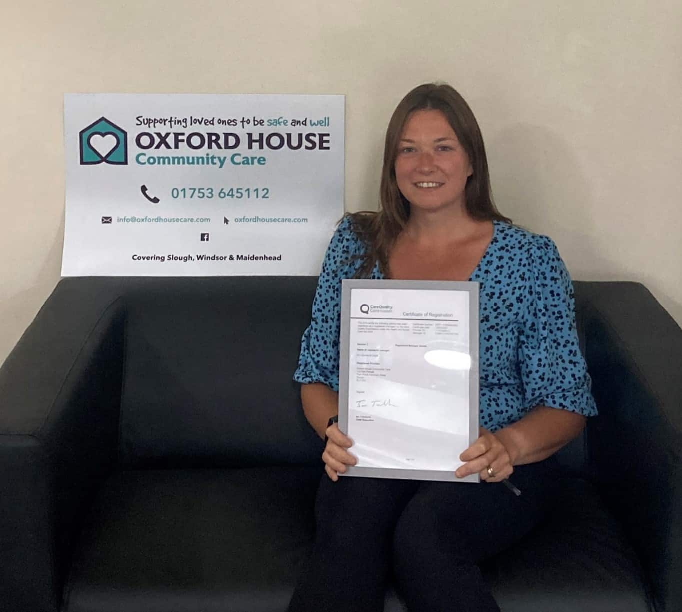 A smiling woman sitting on a couch holding a certificate, with a banner of oxford house community care in the background indicating support for loved ones in slough, windsor, and maidenhead.