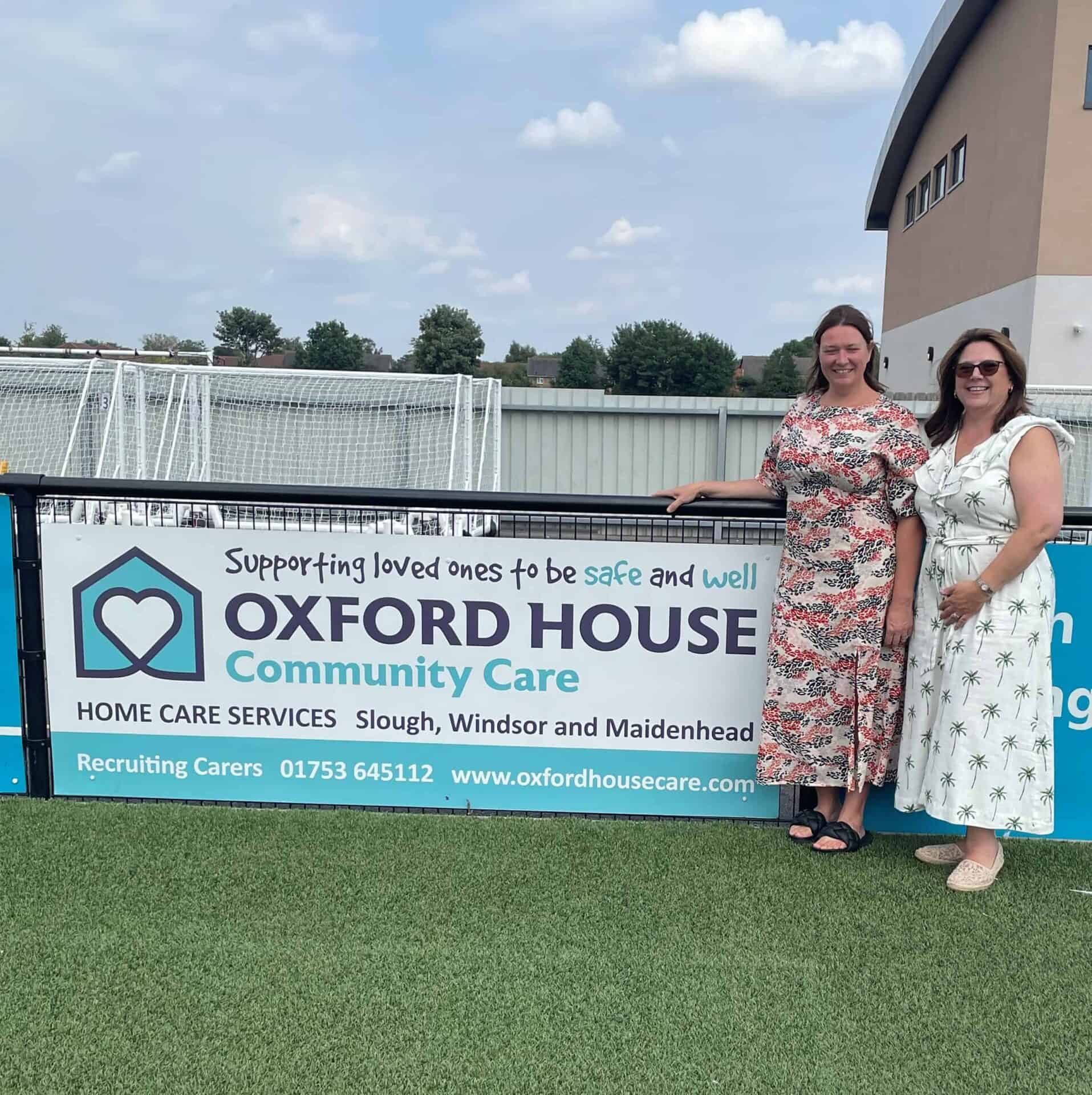 Two smiling women standing in front of a banner for oxford house community care, promoting services for supporting loved ones to be safe and well, with contact information below.