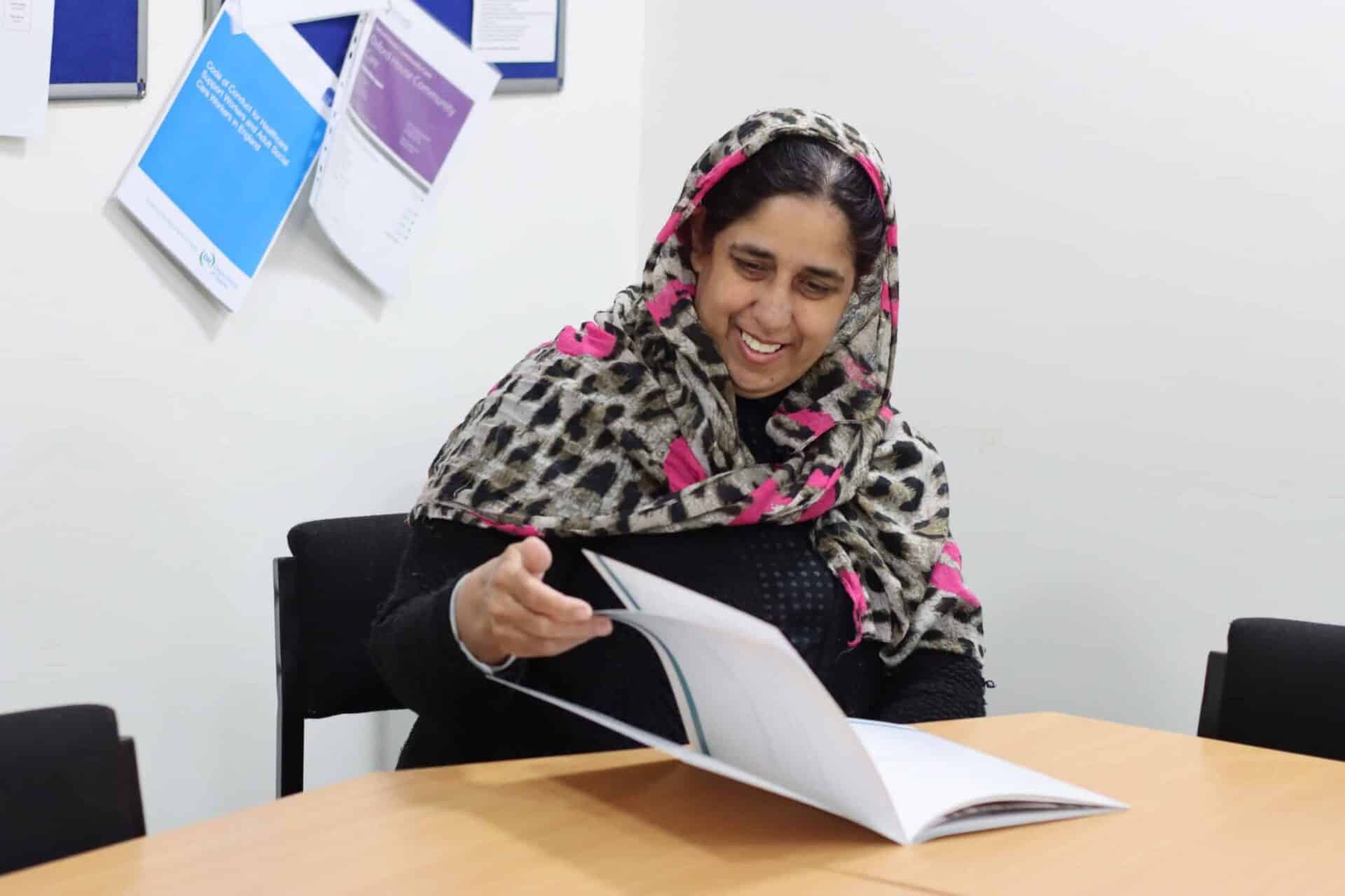 A smiling woman in a patterned headscarf, engaged in reading a report at a conference table.