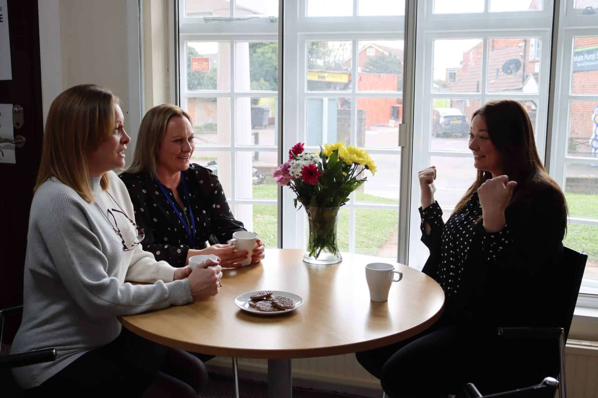 Three women sharing a pleasant conversation over coffee at a round table with a vase of fresh flowers in the centre.