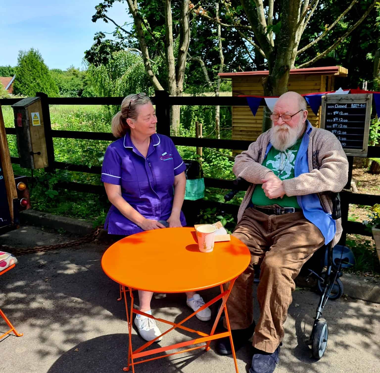 A caregiver in a purple uniform from Oxford House Community Care sits with a smiling elderly man with a white beard in a wheelchair, enjoying a sunny day outdoors with an orange table between them, a beverage on top