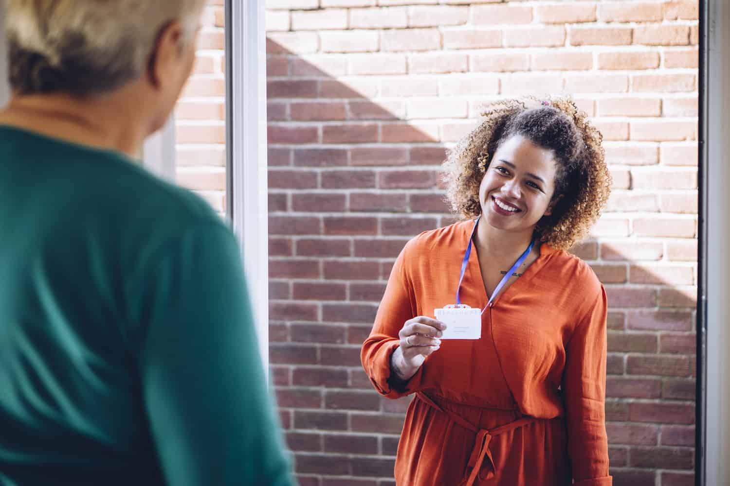 A smiling professional woman with an ID badge welcomes someone at the door for home care services.
