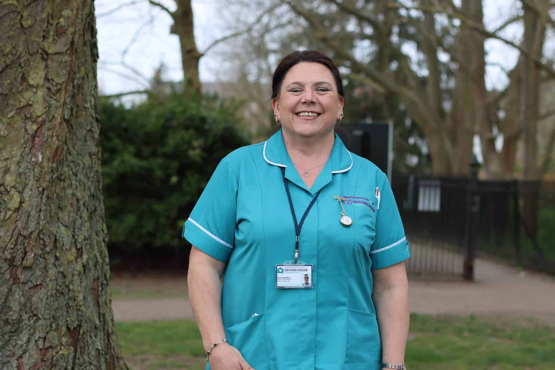 A smiling healthcare professional wearing scrubs and a badge from Oxford House Community Care stands confidently in a park-like setting.