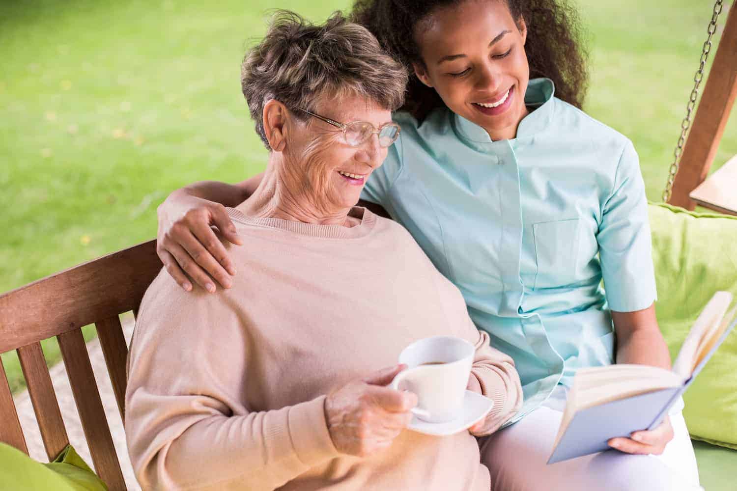 Sharing stories and warm smiles: a young carer enjoys a book with an elderly woman on a relaxing day, as they bond over a cup of tea on a cozy garden swing.