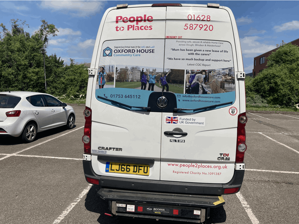 A white minibus with "People 2 Places" and contact information alongside an advert for Oxford House Community Care displayed on the back, parked in a lot with other cars on a sunny day.