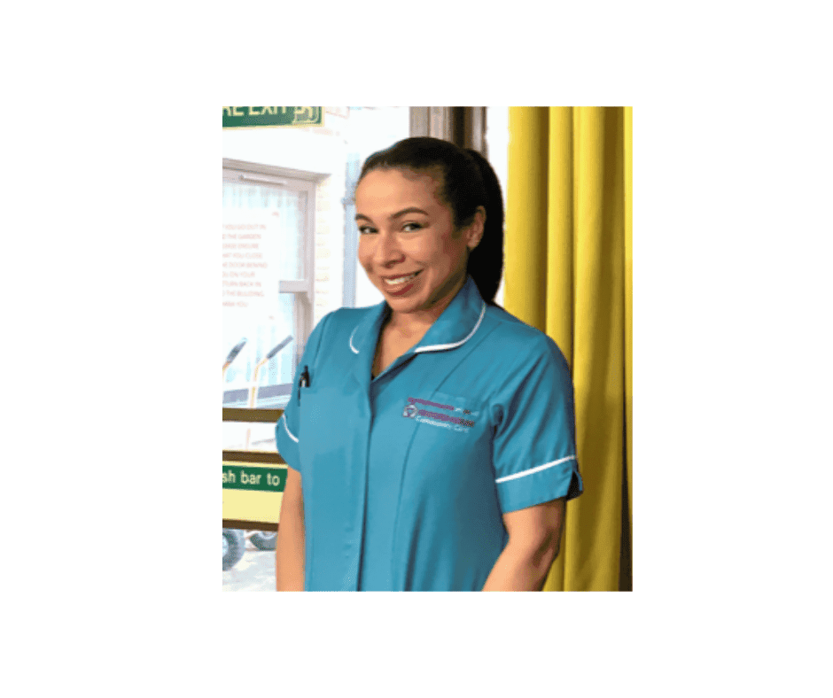 A smiling woman in a teal uniform standing indoors, with a friendly and approachable demeanour.