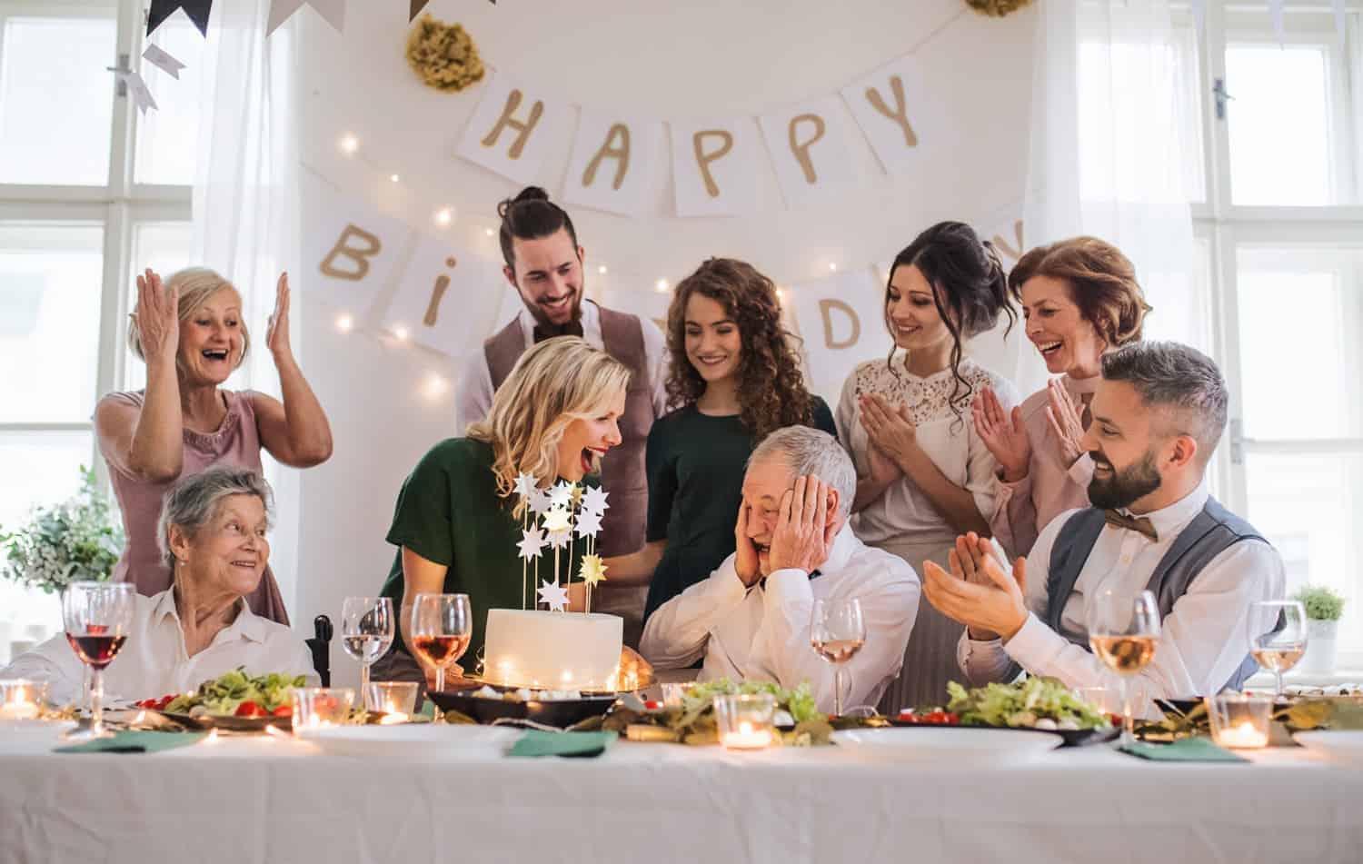 A joyful family gathering to celebrate a senior man's birthday, with loved ones cheering and smiling around a table decorated with flowers and candles, under a banner reading "happy birthday".