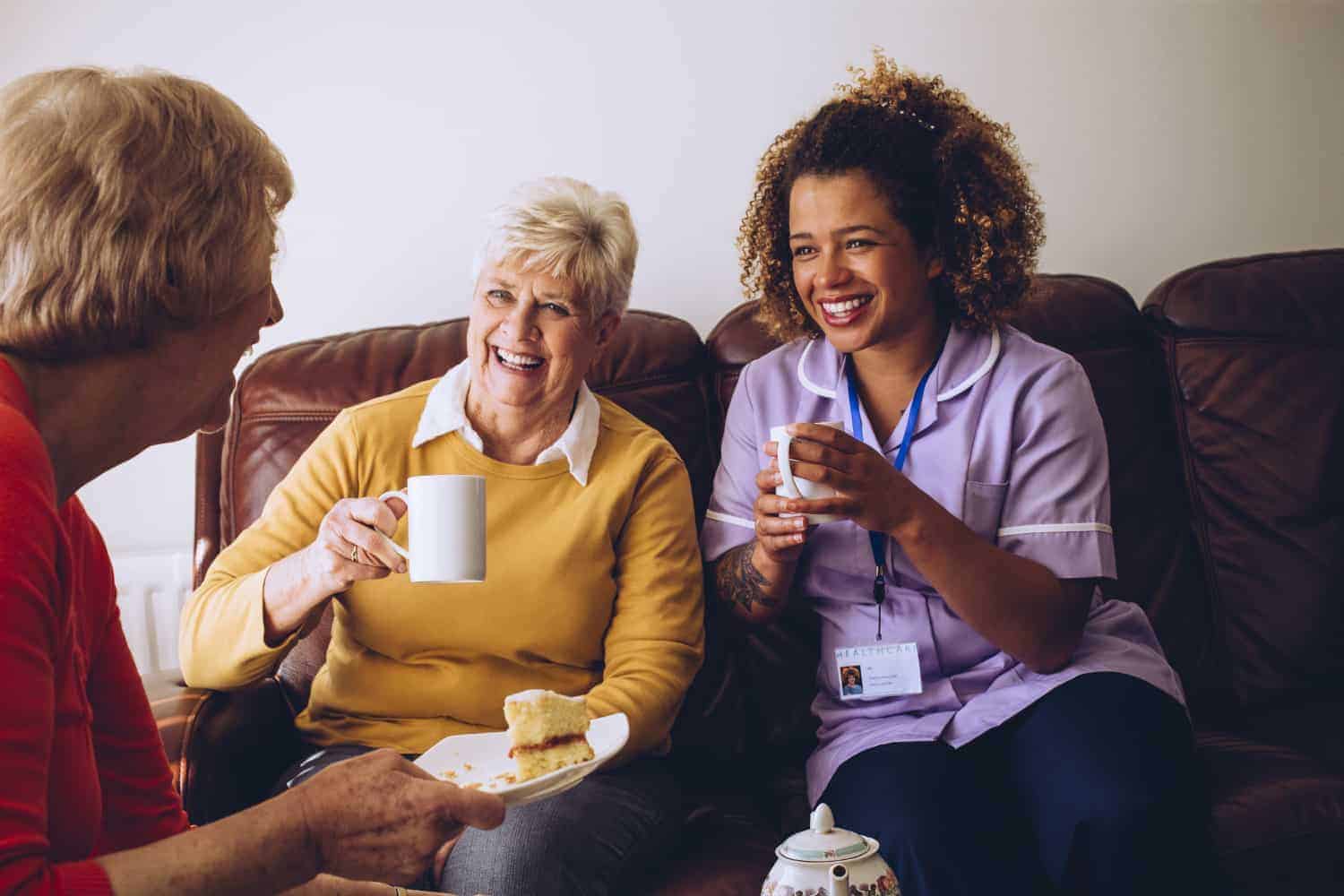 Three women sharing a joyful moment over tea and snacks, with the youngest wearing a nurse's uniform, suggesting a warm home care provider-patient relationship.