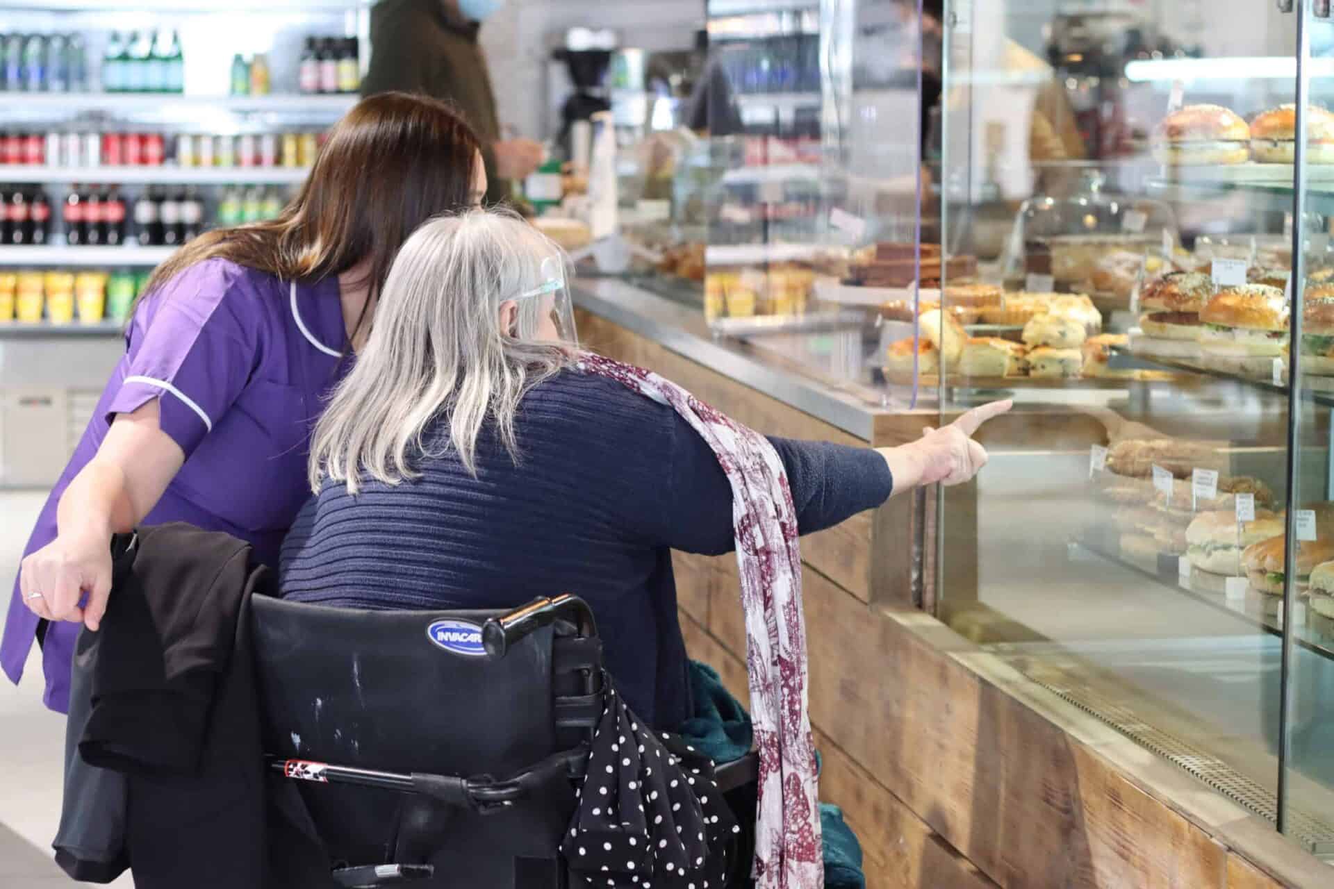 An elderly woman in a wheelchair is assisted by another woman as they look at a display of pastries in a store, with the seated woman pointing at something that caught her interest.