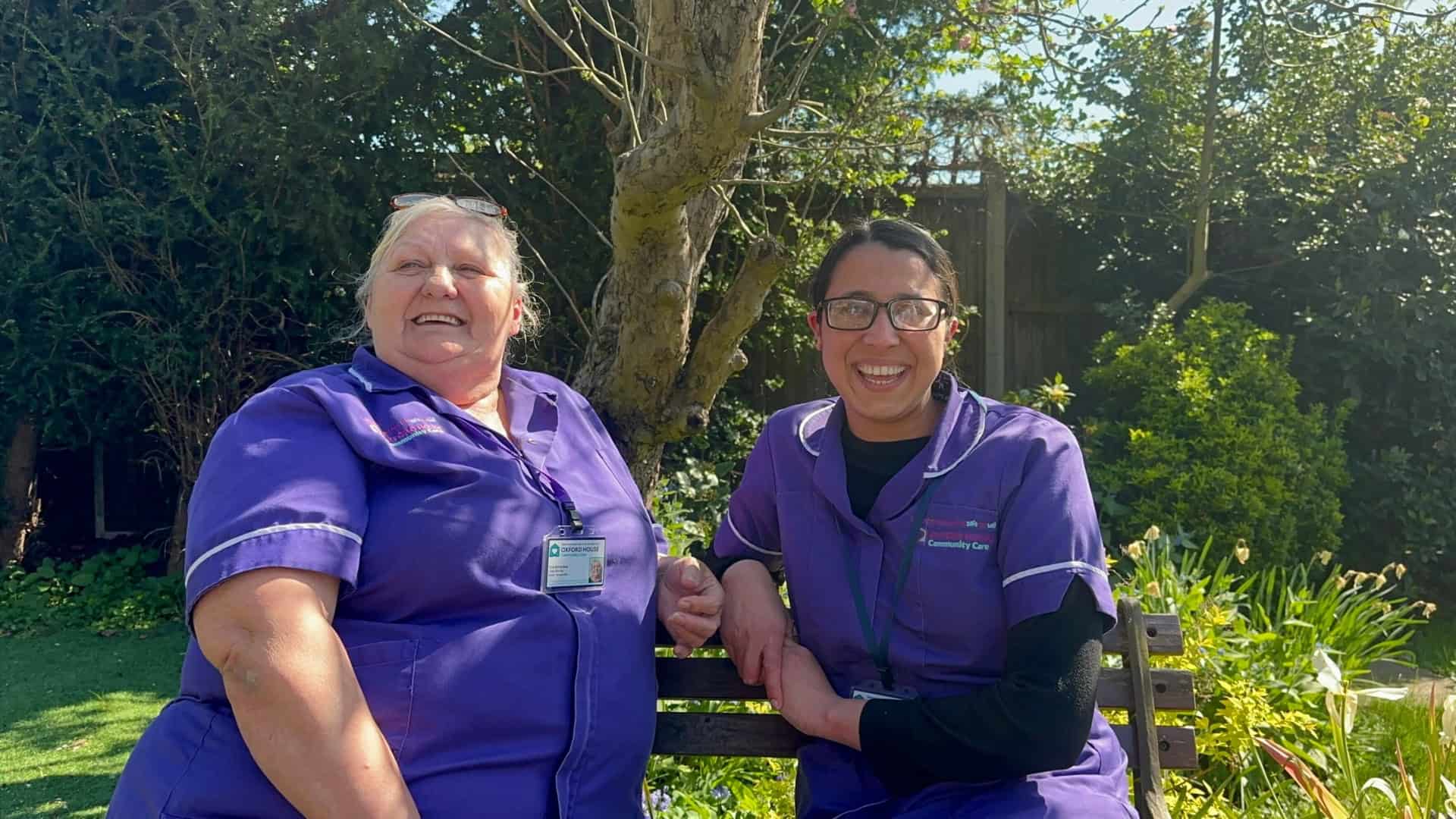 Two women wearing purple uniforms and name badges sit on a bench outdoors, smiling and laughing. A tree and greenery are in the background, and sunlight brightens the scene.
