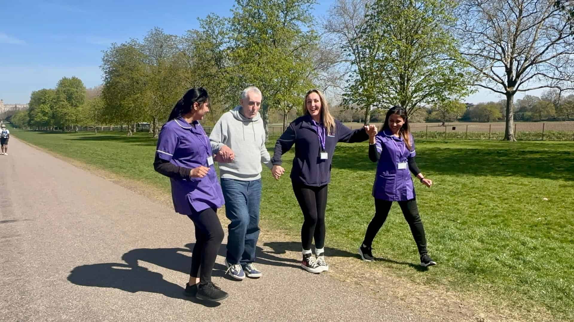 Three women in purple uniforms walk and hold hands with an older man in a park on a sunny day. The group appears cheerful, surrounded by green grass and trees under a clear blue sky.