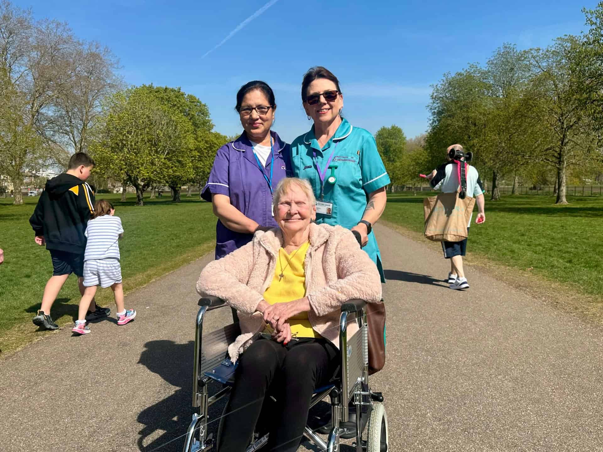 Two nurses stand smiling behind an older woman seated in a wheelchair on a sunny park path. Other people, including children, walk and play in the background among green trees.