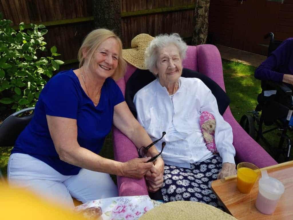 Two cheerful women enjoying a sunny day outdoors, with one sitting in a wheelchair, both wearing hats and sharing a warm moment together, highlighting the personalized care between them.