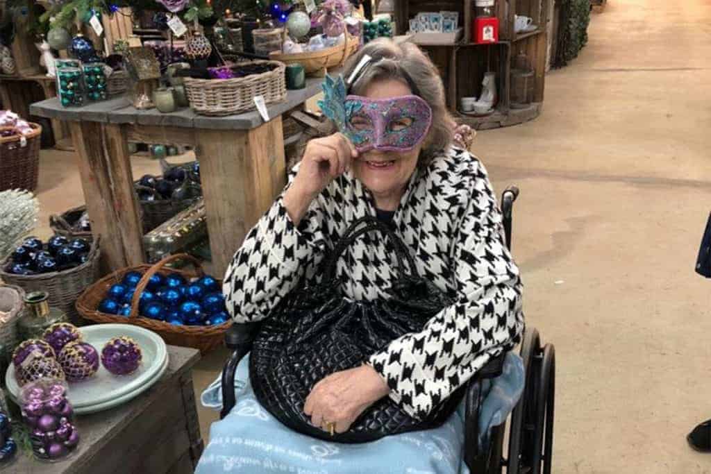 An elderly woman in a wheelchair trying on a colorful masquerade mask in a Berkshire care home, surrounded by various home decor items.