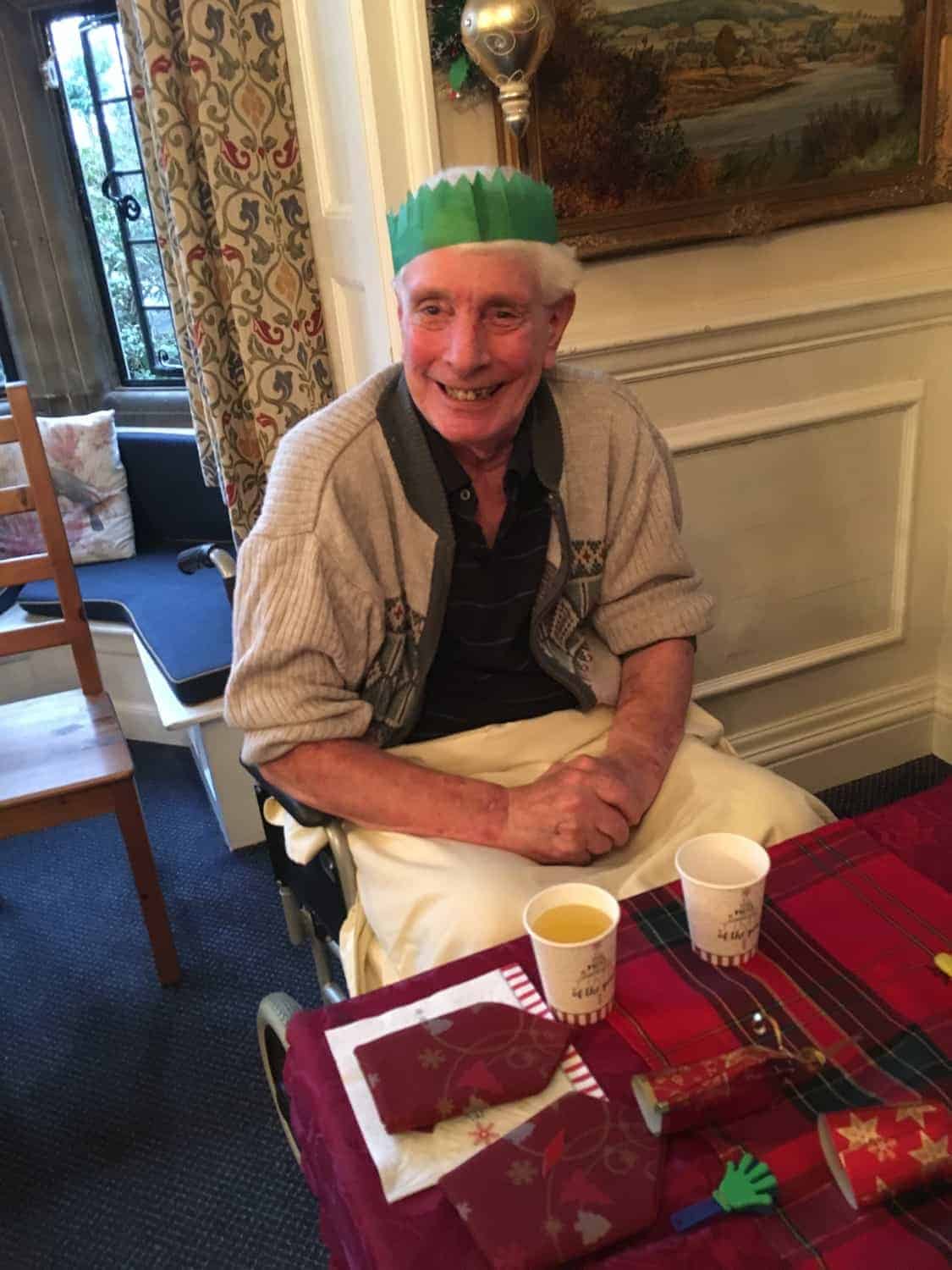 An elderly gentleman wearing a green paper crown and a warm smile, seated at a table with festive decorations in a gallery, celebrating the holiday spirit.