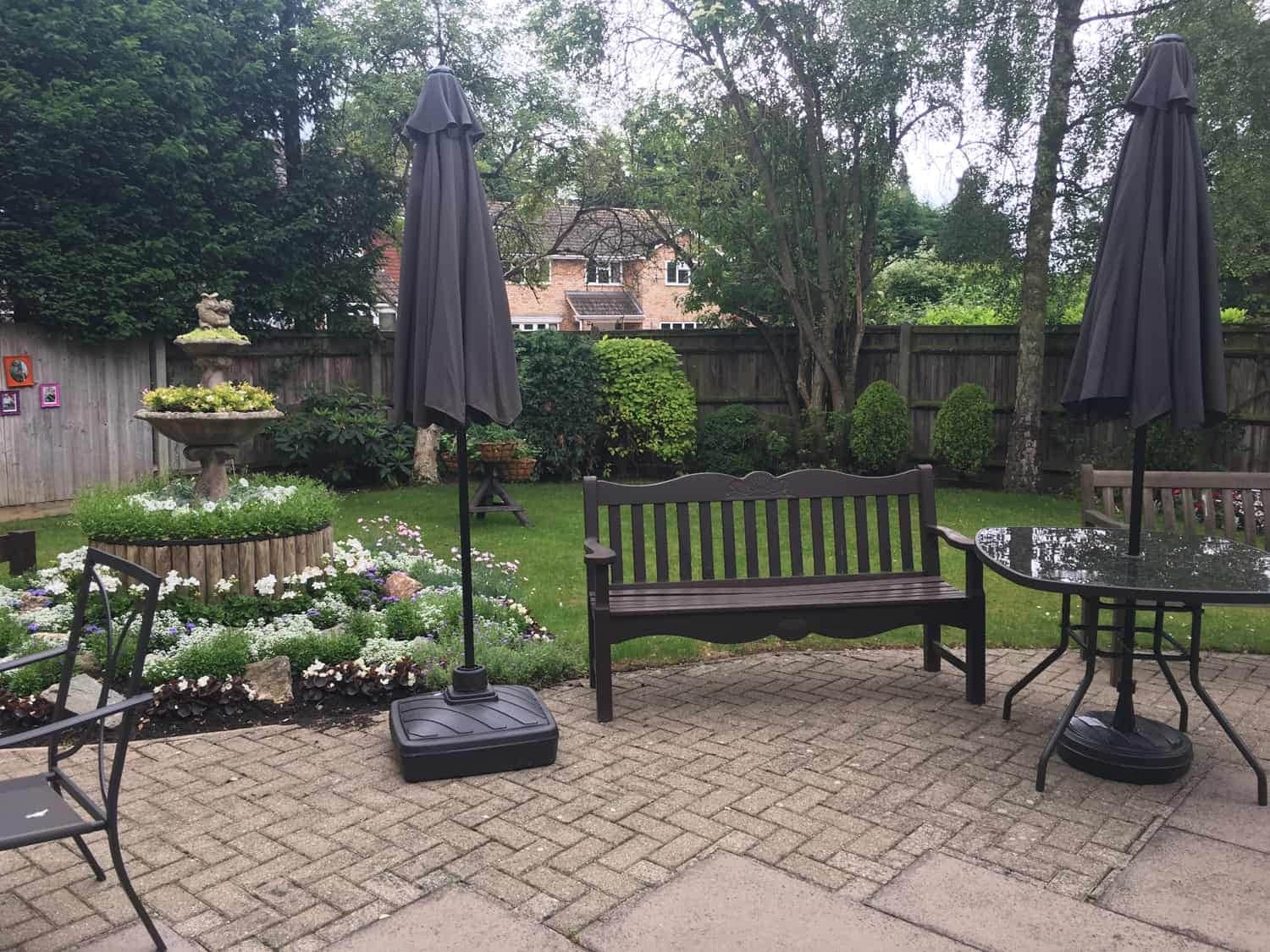 A tranquil garden patio with closed umbrellas, a wooden bench, and a table surrounded by blooming flowers, lush greenery, and contemporary art exhibits.