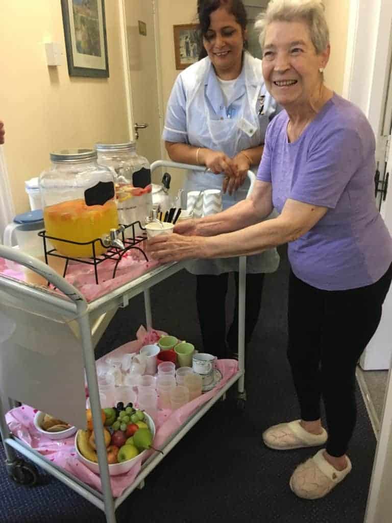 A cheerful elderly woman enjoys a refreshing beverage from a beverage cart, served by a smiling healthcare worker in a corridor, promoting her health and wellbeing.