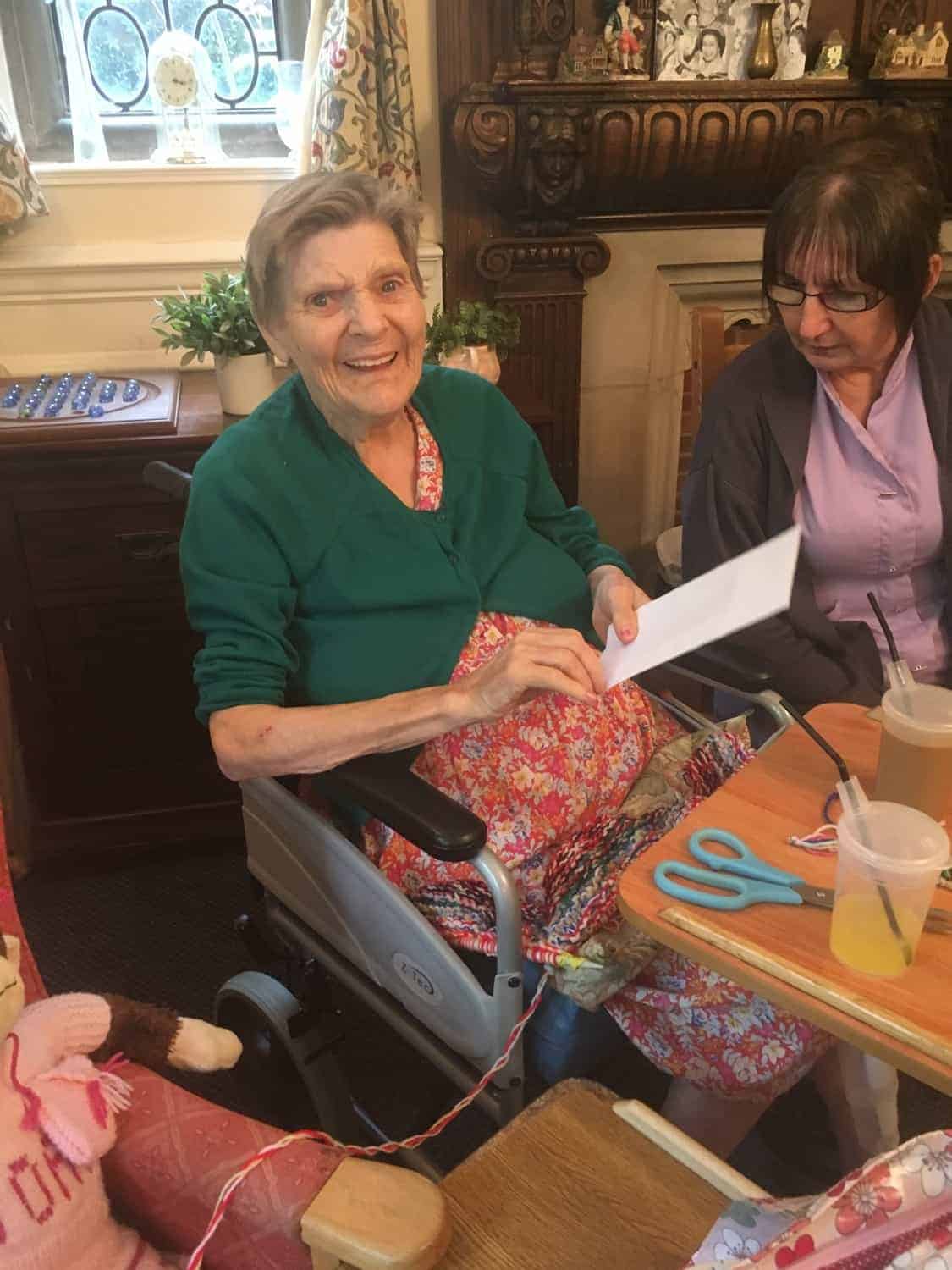 An elderly lady in a wheelchair with a bright green cardigan and floral dress is smiling brightly as she looks at the camera, whilst a woman sitting next to her assists her with some papers. They appear