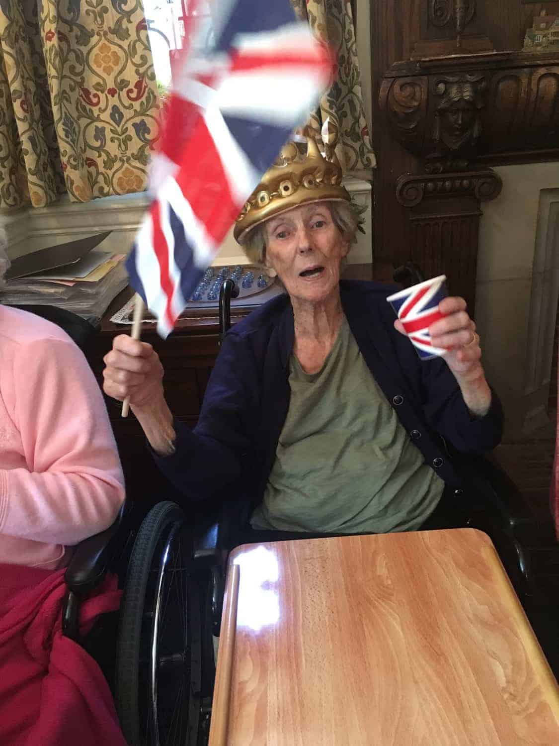 An elderly individual cheerfully waving a British flag, seated in a wheelchair in a gallery, with a joyous expression and perhaps celebrating a national event or showing patriotic spirit.