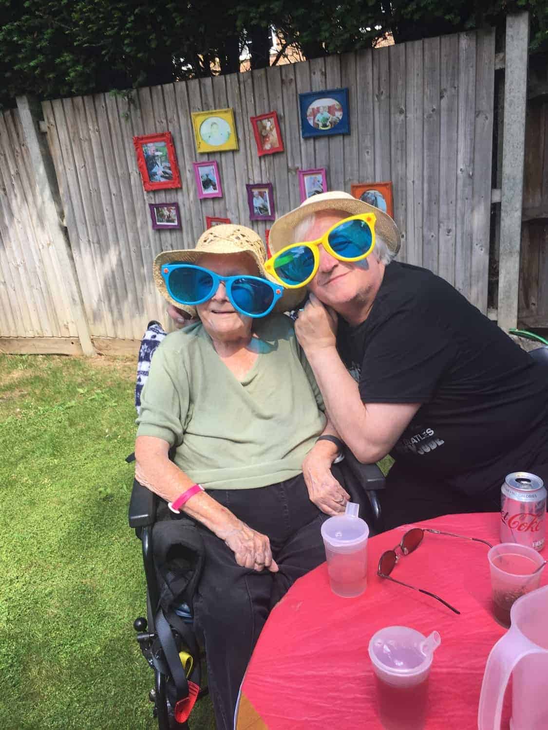 Two individuals at an outdoor gallery gathering, sporting large playful sunglasses and hats, sharing a happy moment together.
