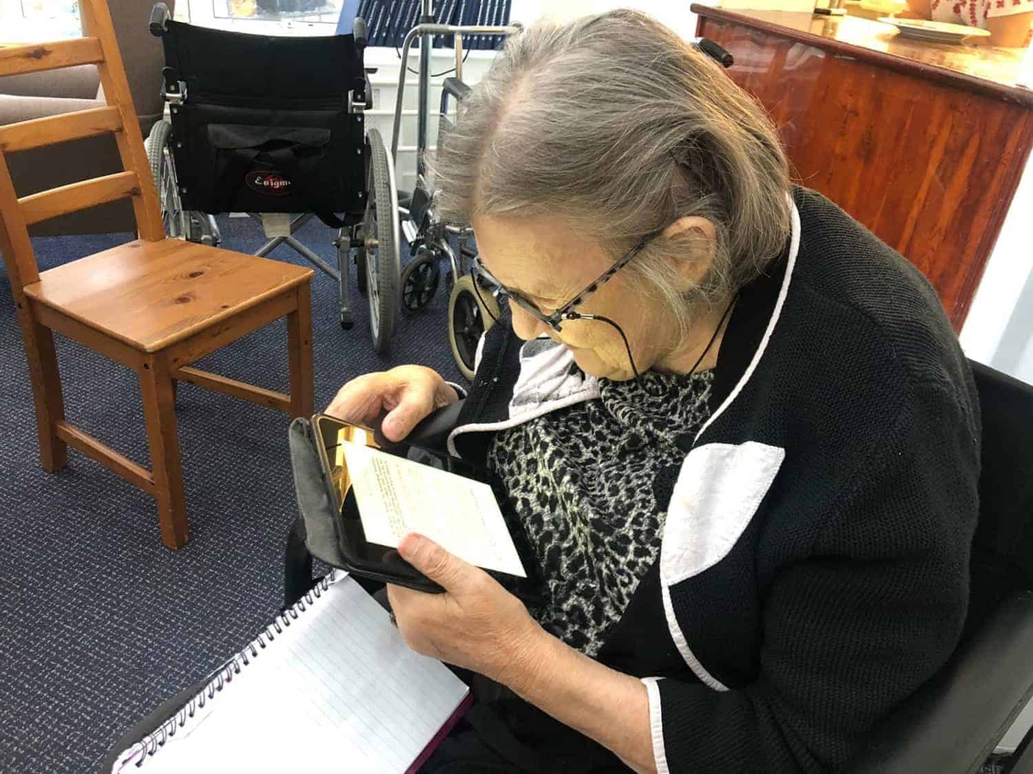 An elderly woman with glasses is engaged in examining artwork content on a smartphone, seated next to a notebook and a wheelchair in an art gallery setting.