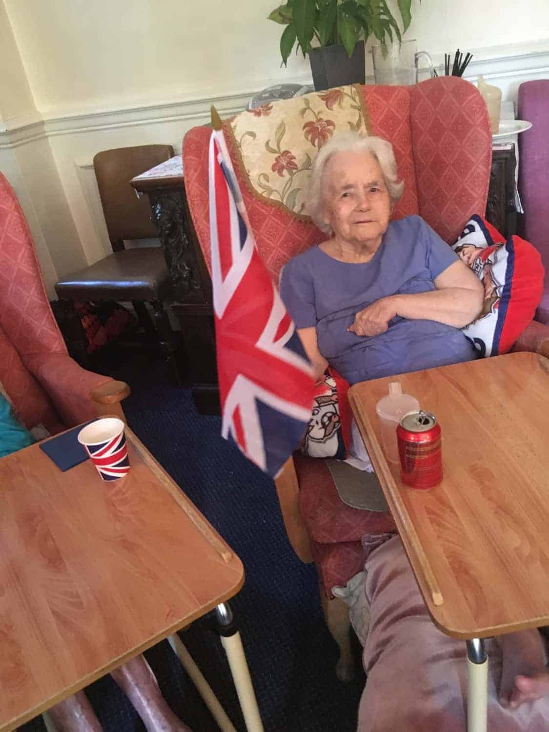 An elderly lady sitting comfortably in a chair, with a British flag displayed next to her in an art gallery, exuding a sense of national pride and contentment.