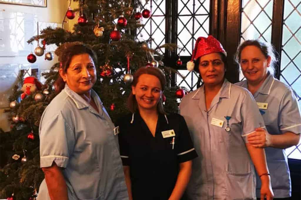 Four healthcare professionals in uniform smiling for a festive photo in front of a decorated Christmas tree in a family-run home, with one wearing a Santa hat to celebrate the holiday spirit.