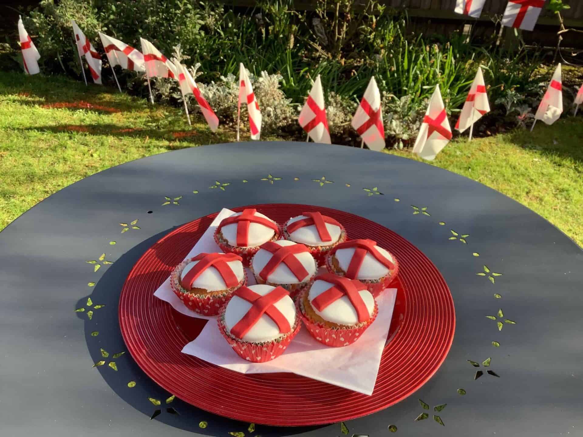 A festive outdoor table setting featuring a plate of cupcakes decorated with red and white icing to resemble the English flag, surrounded by a garden adorned with matching flag bunting, celebrating community events or national holiday.