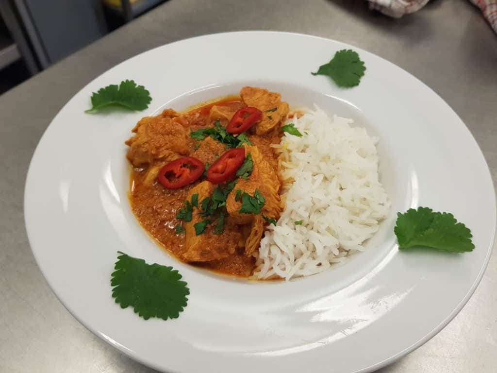 A plate of homemade flavourful chicken curry garnished with fresh coriander and slices of red chilli, accompanied by a side of steamed white rice.