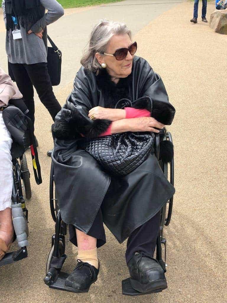 An elderly lady in sunglasses and a black outfit, with a fur stole around her shoulders, calmly sits in a wheelchair outdoors at an art gallery, exuding a sense of elegance and grace.
