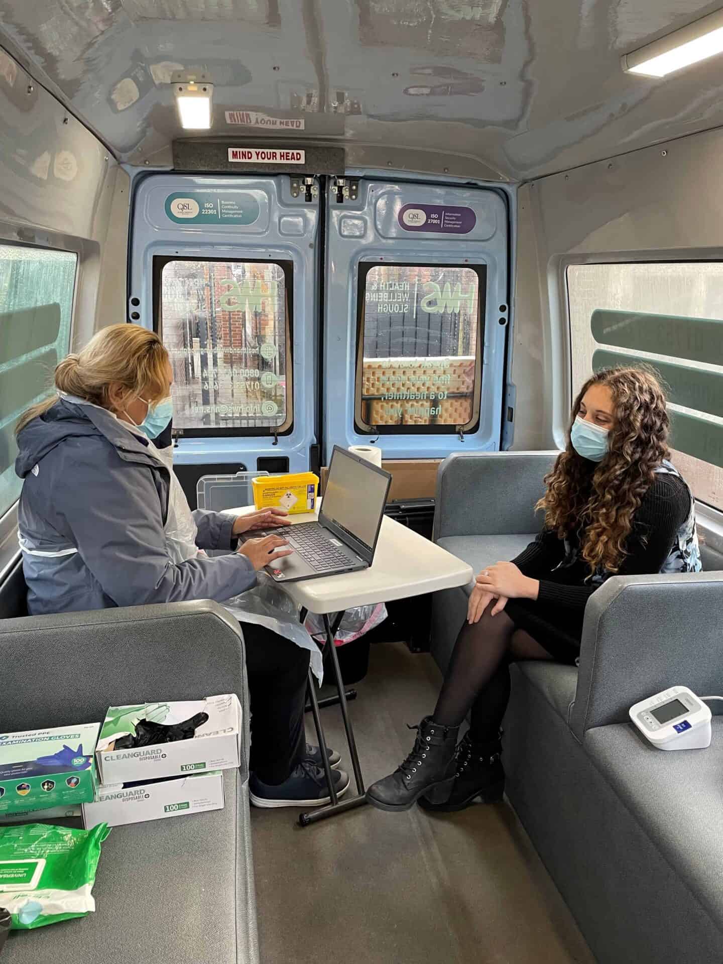 A healthcare worker and a patient inside a mobile clinic in Slough, with the patient about to receive medical attention or consultation while wearing protective masks.