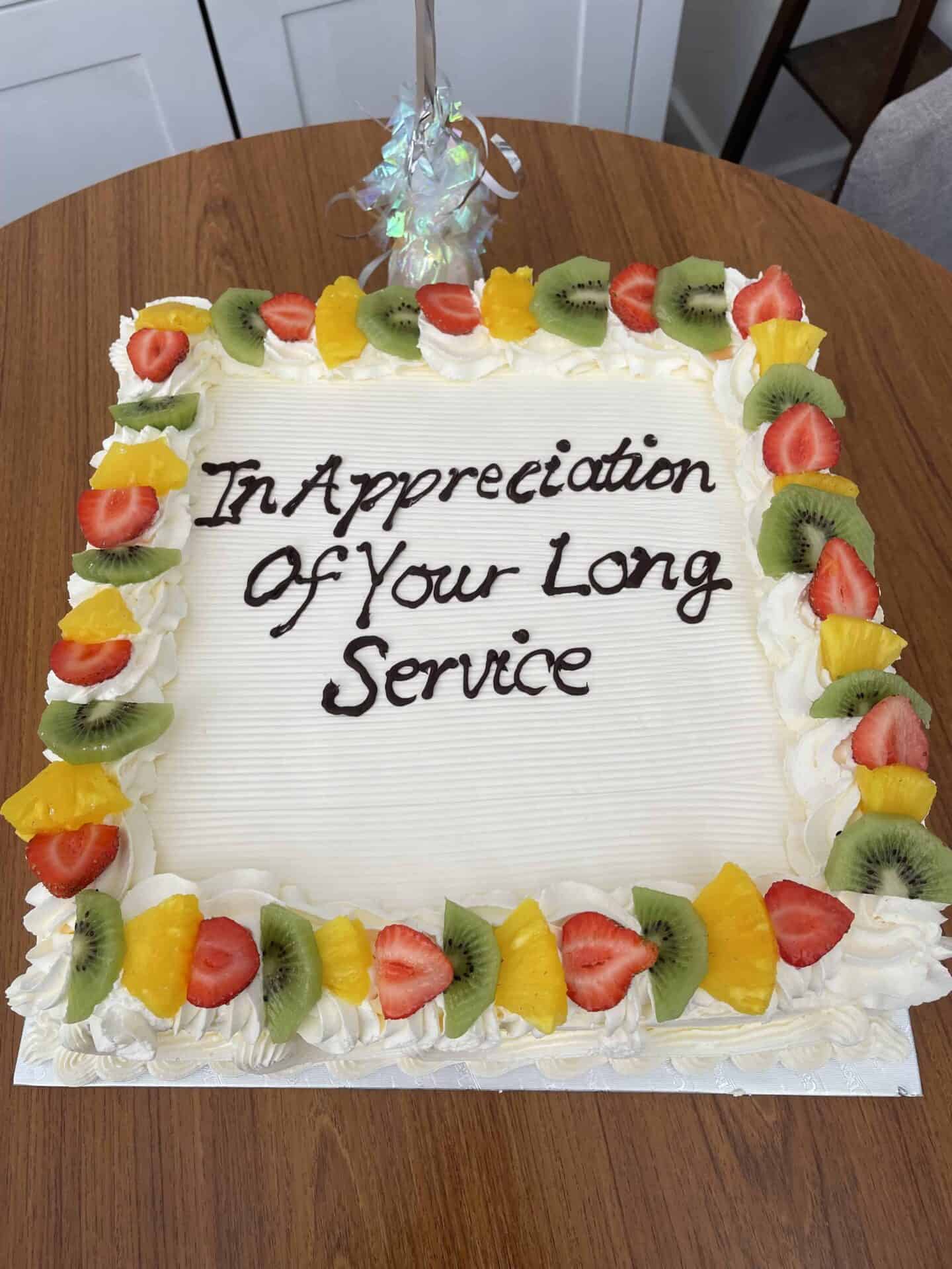 A large rectangular celebration cake with the message "in appreciation of your long service" written in chocolate frosting, decorated with whipped cream dollops and fresh fruit slices including strawberries, kiwis, and oranges.