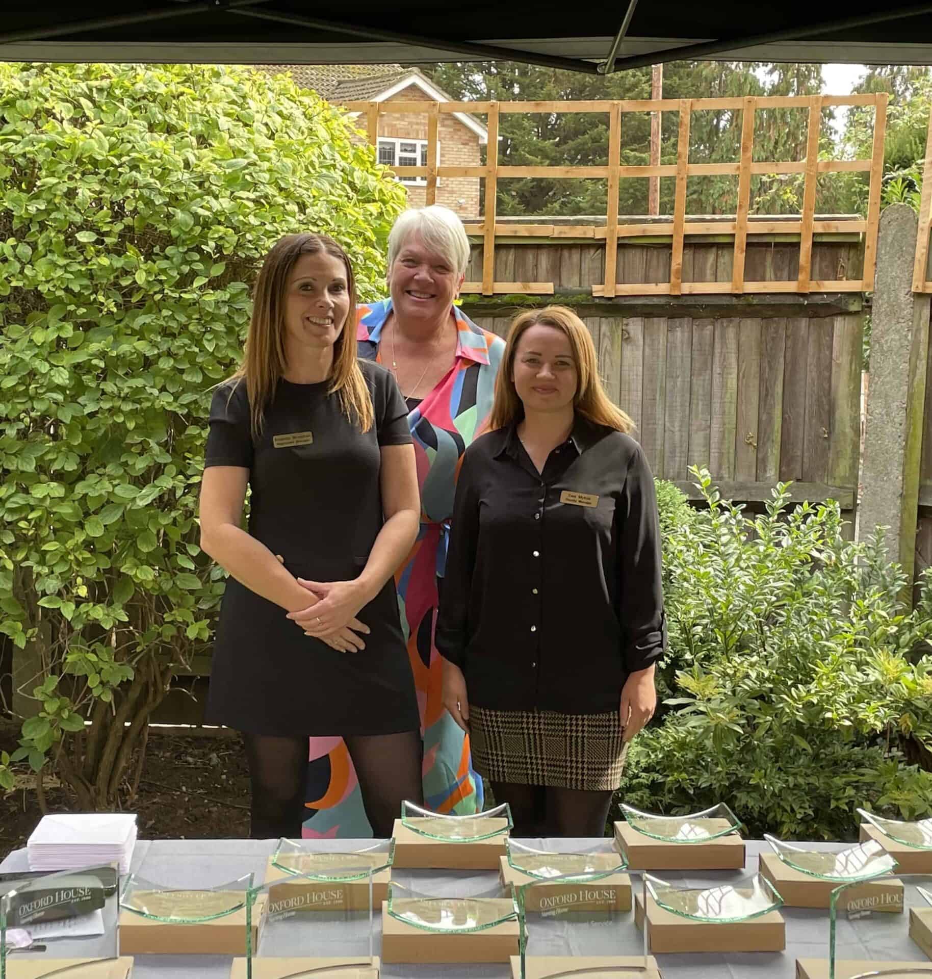 Three women standing behind a table with neatly arranged boxes, smiling and posing for a photo at what appears to be an outdoor event or gathering.