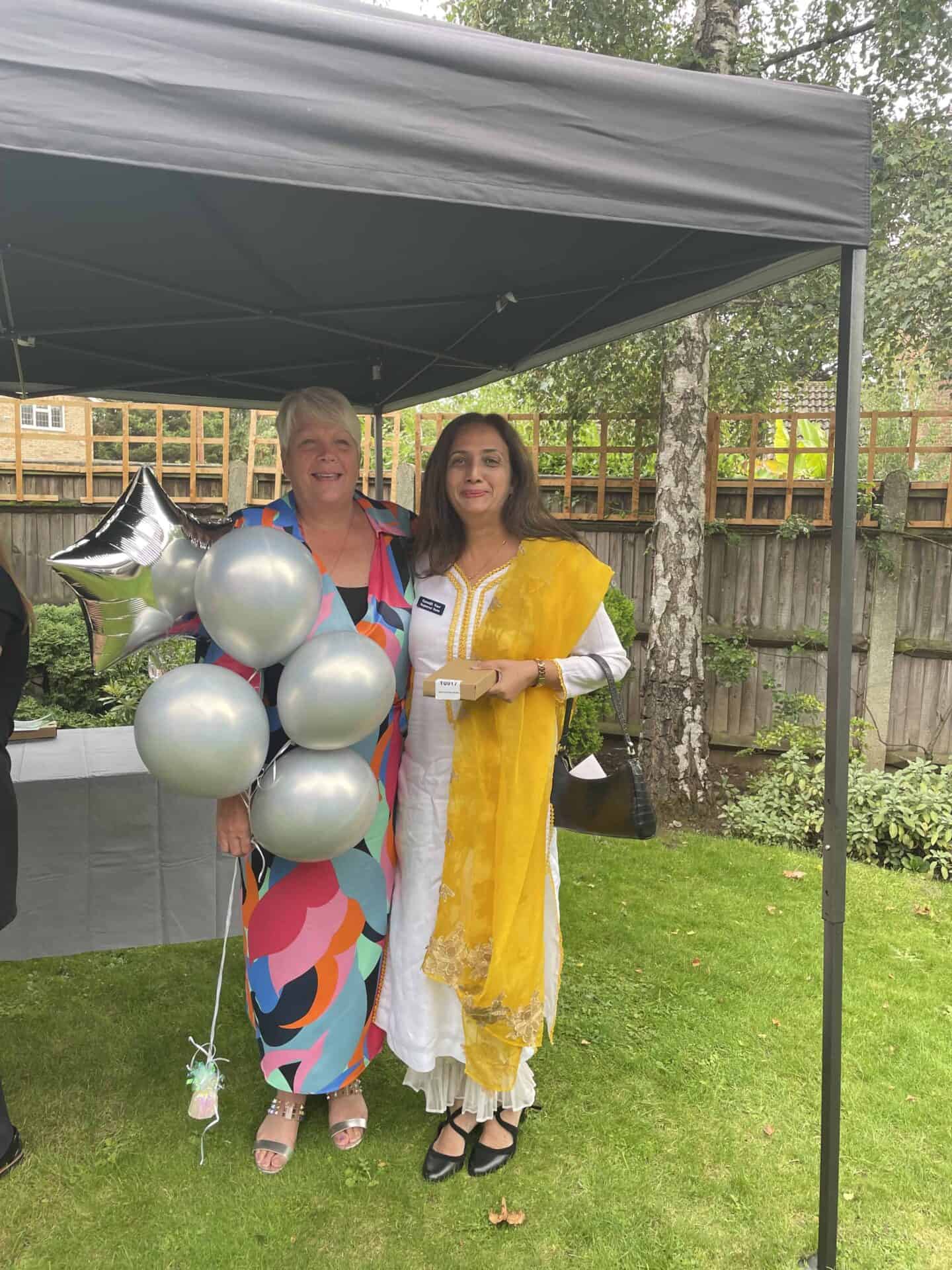 Two smiling women at an outdoor event, one holding a bunch of silver balloons, both dressed for a festive occasion, with a garden and a canopy in the background.