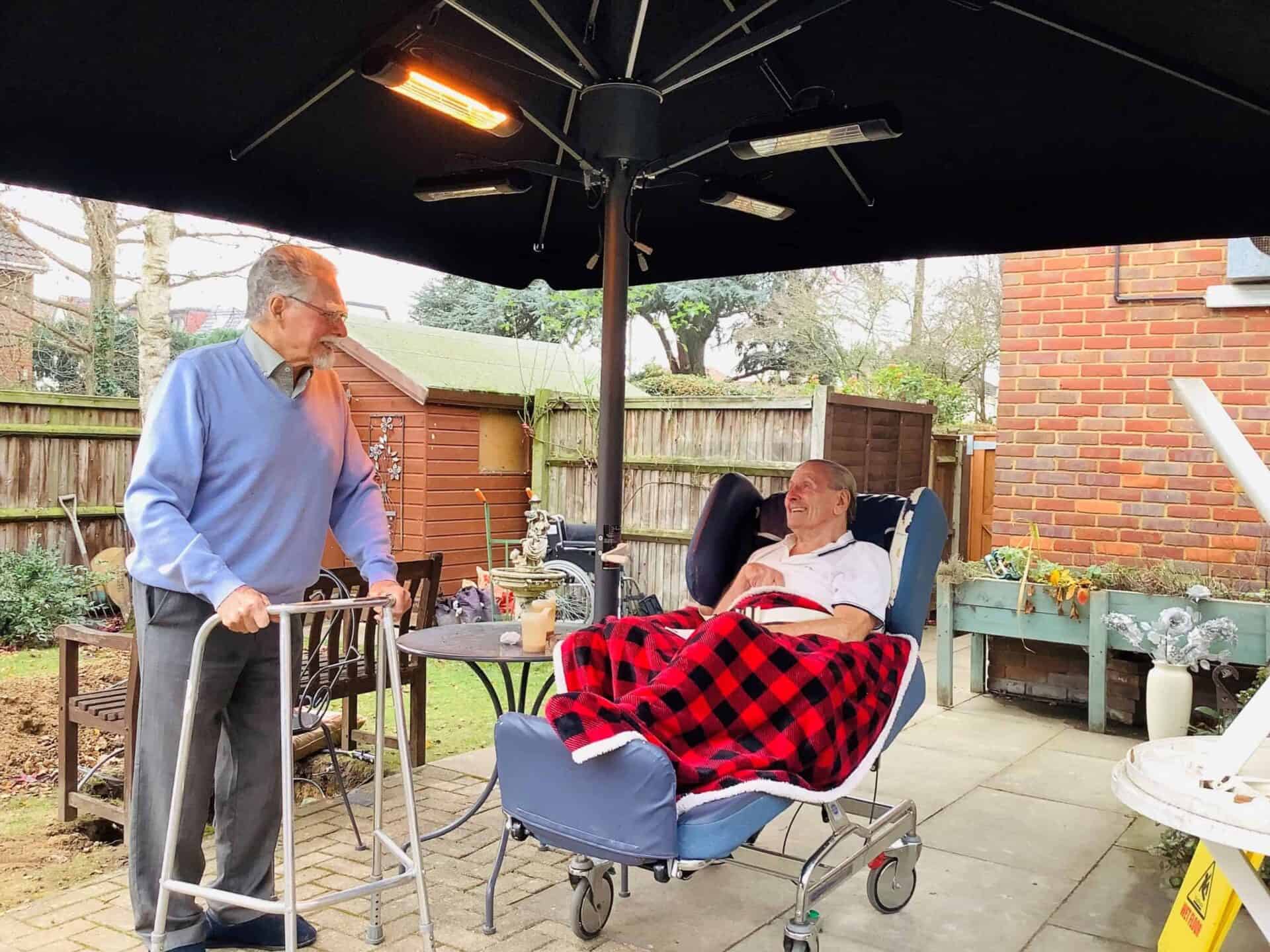 Two elderly gentlemen enjoying a conversation outdoors, one standing with the help of a walking stick and the other relaxing in a reclining wheelchair, under the shade of a large umbrella with heaters, surrounded by the