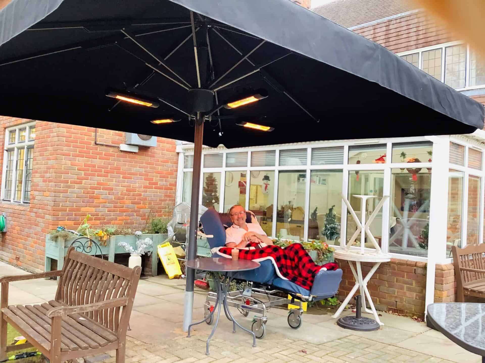 Relaxed outdoor lounging: a person wrapped in a cozy blanket reclines on a patio chair under a large umbrella with heating lamps, enjoying a tranquil moment outside in an artistically landscaped garden.