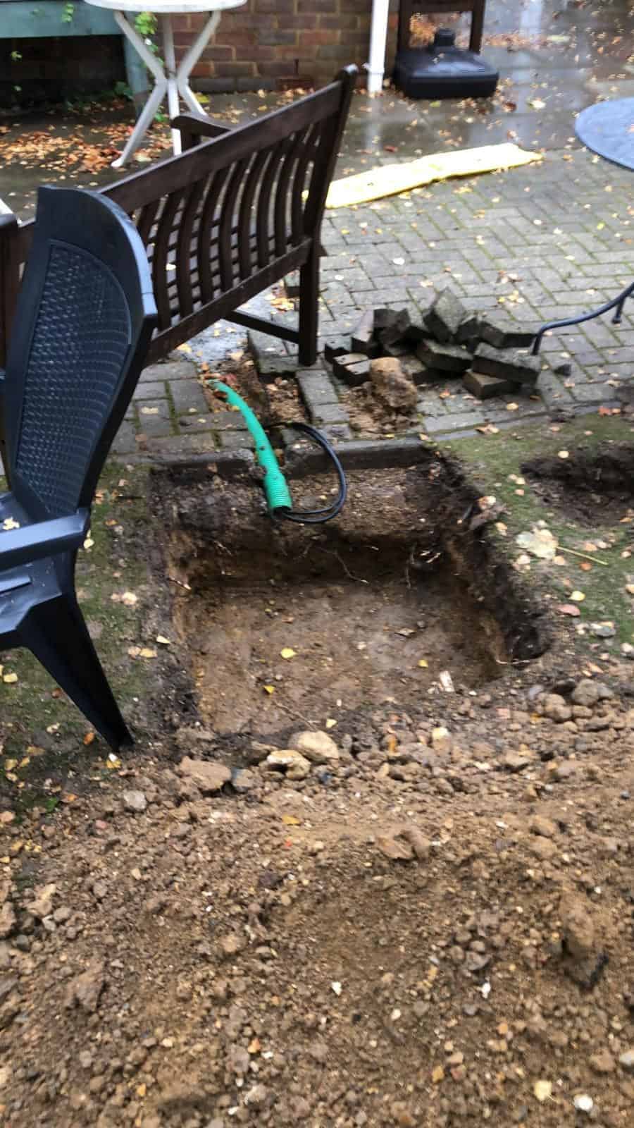 A partially excavated square trench revealing underground infrastructure next to uprooted paving stones and disarranged garden furniture, suggesting recent groundwork for a new garden project.