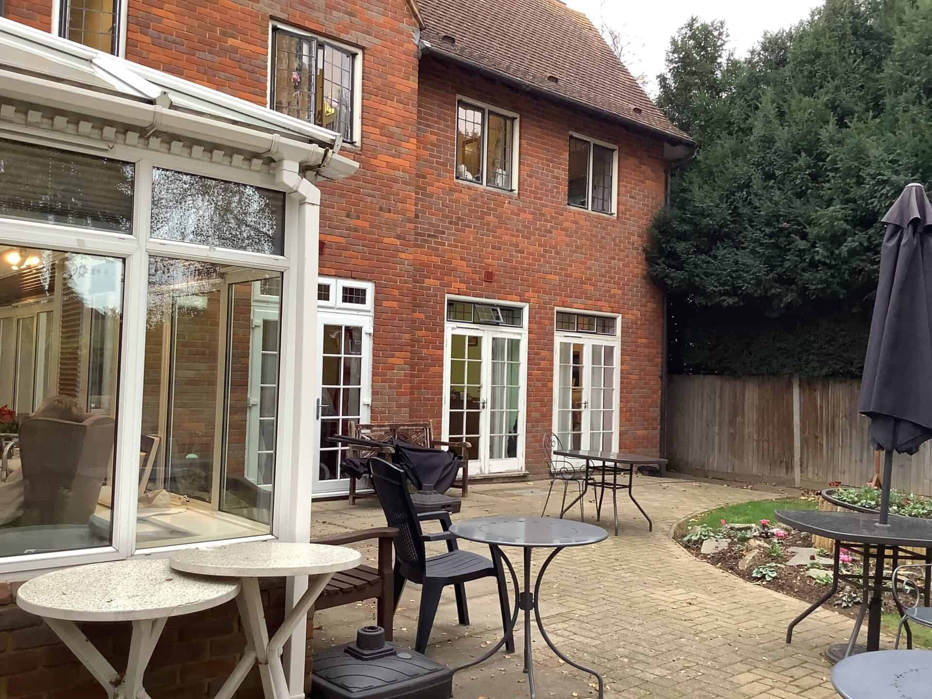 Brick house with a garden design featuring a conservatory extension and a cozy patio area furnished with tables and chairs, set against a backdrop of lush greenery.