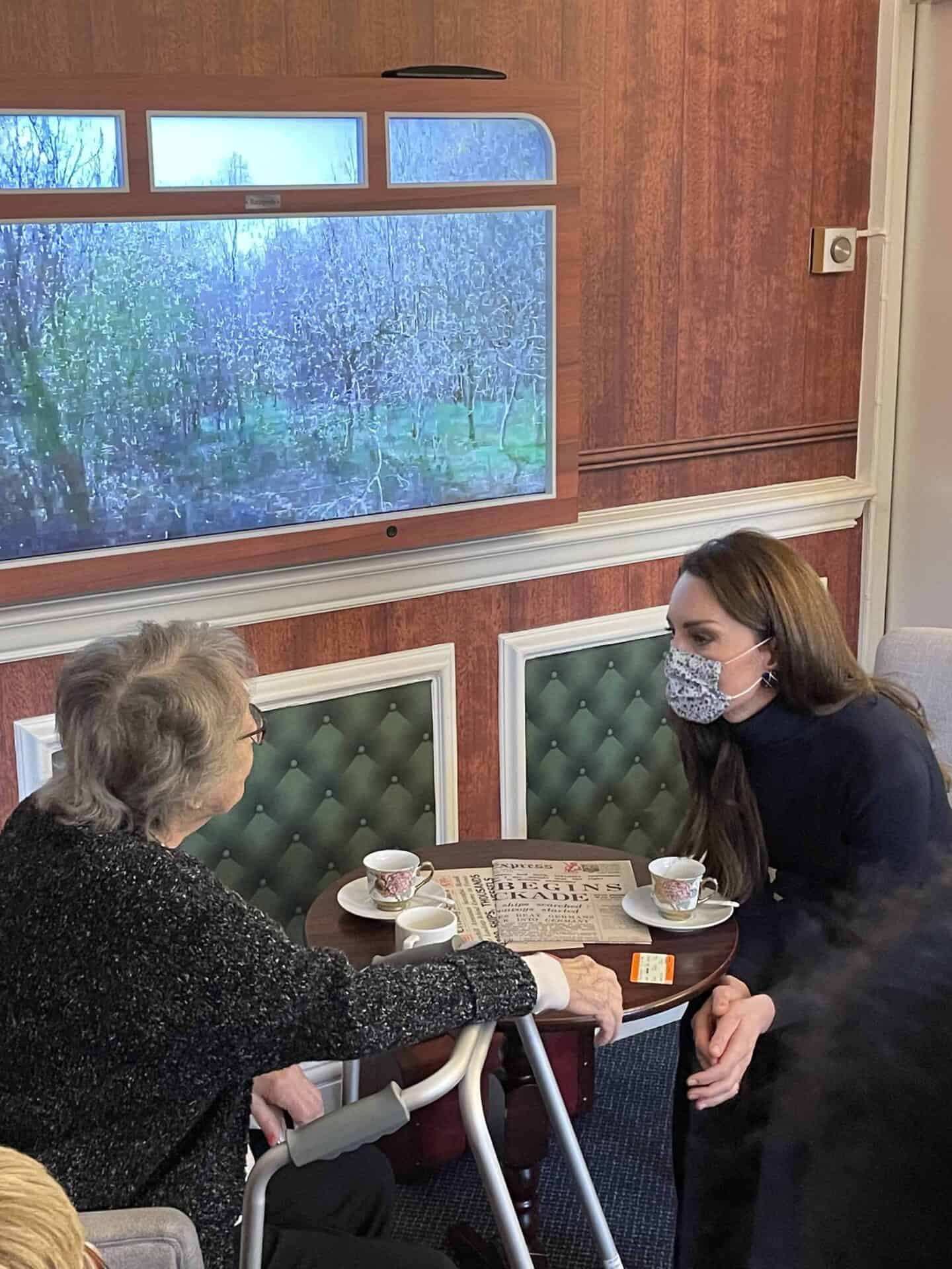The Princess of Wales in a protective face mask attentively listens to an elderly lady seated with a walker as they sit across from each other at a table with a large crossword puzzle, featuring an interactive feature.