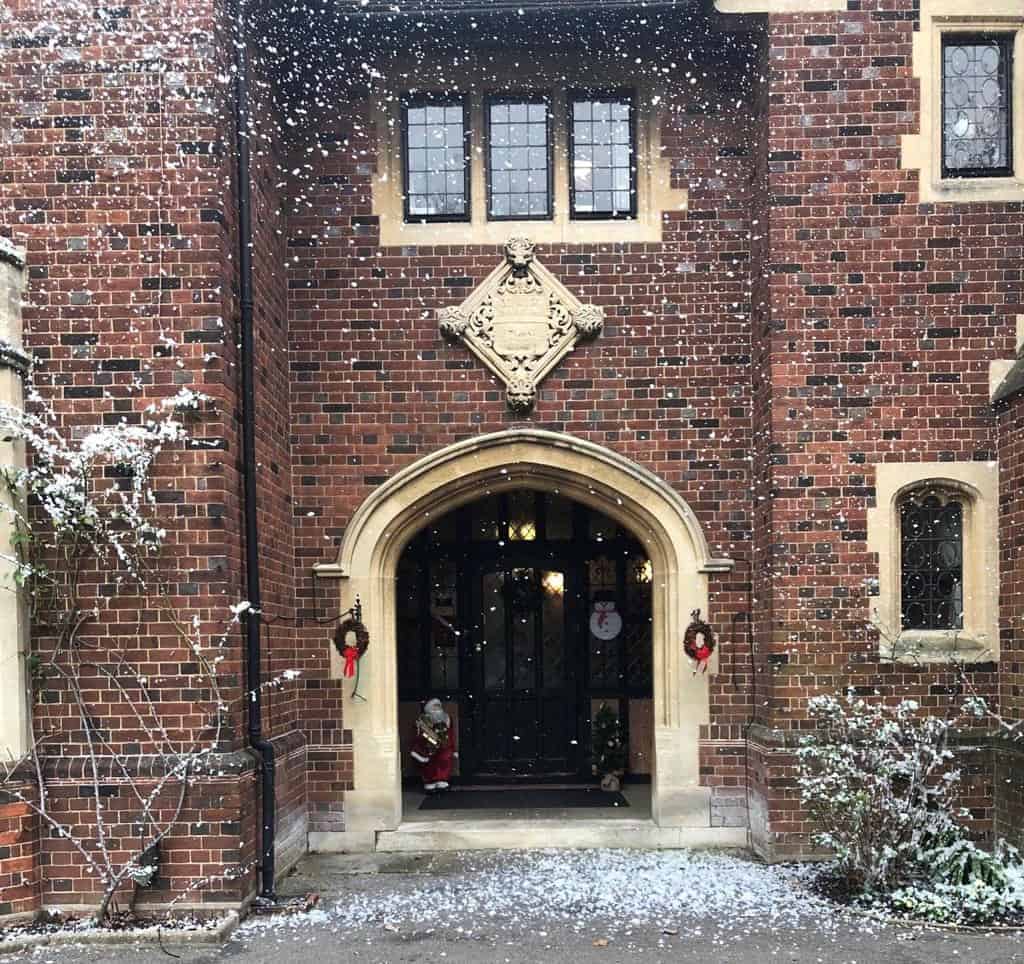 An enchanting winter scene at a traditional brick building with an arched doorway, adorned with festive wreaths and a dusting of freshly falling snow, evoking the warmth of the holiday season, now