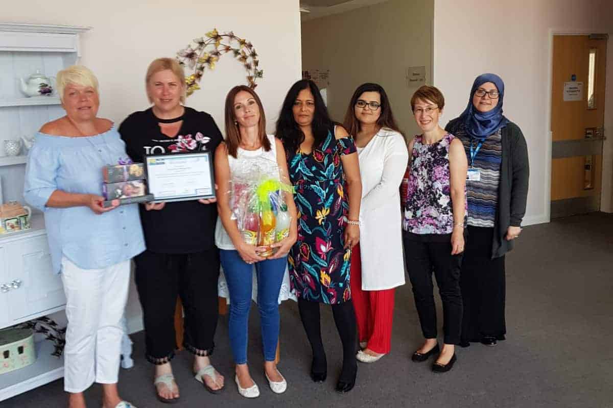 A diverse group of seven women posing for a photo indoors, with some holding awards and gifts, suggesting a celebration of achievement or recognition within an office or community setting, indicative of favorable reviews.