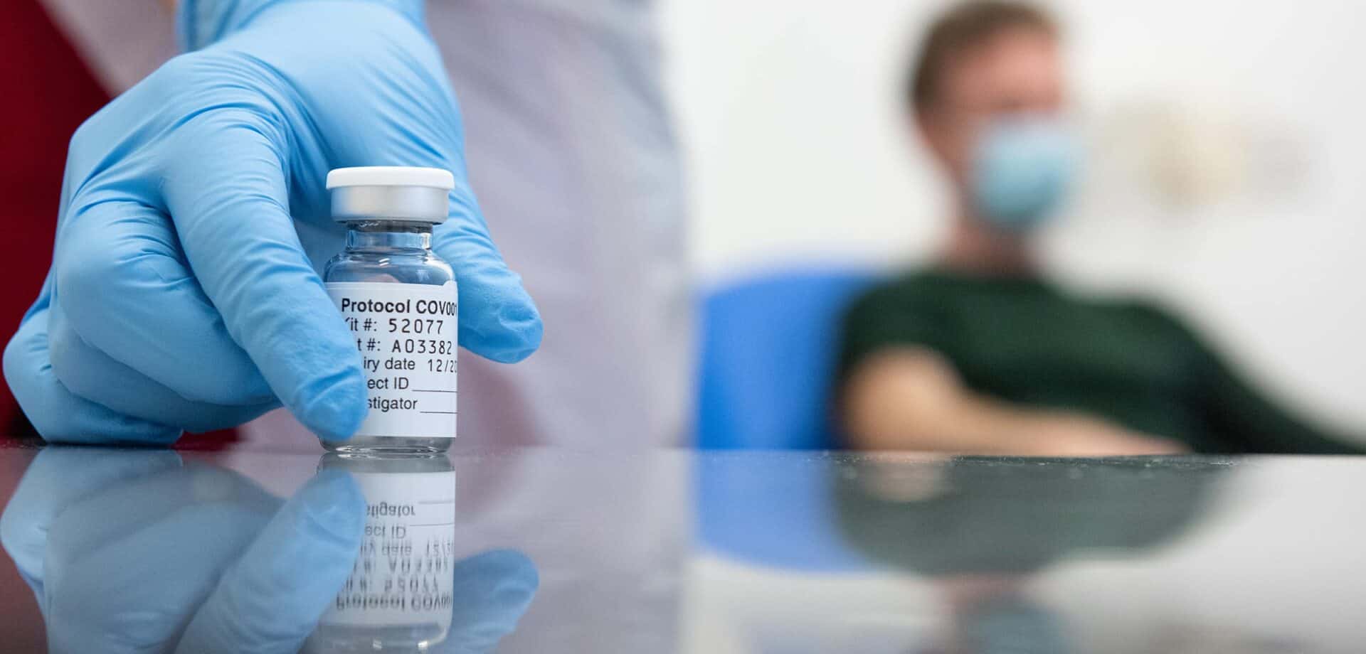 Healthcare professional in blue gloves holding a vial of the COVID-19 vaccine with a patient wearing a mask in the background.